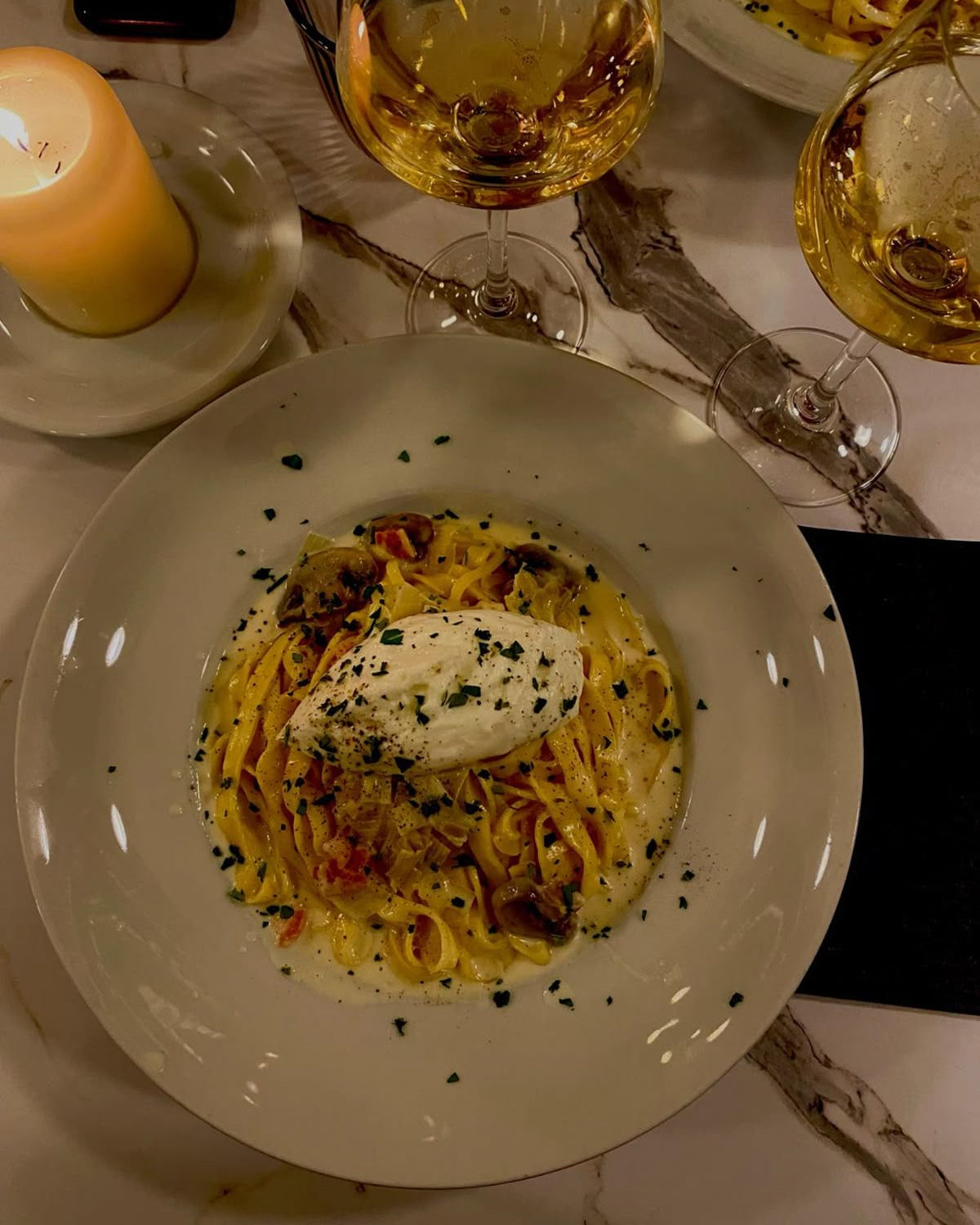 Plate of pasta with a side of wine glasses and a candle on a table.