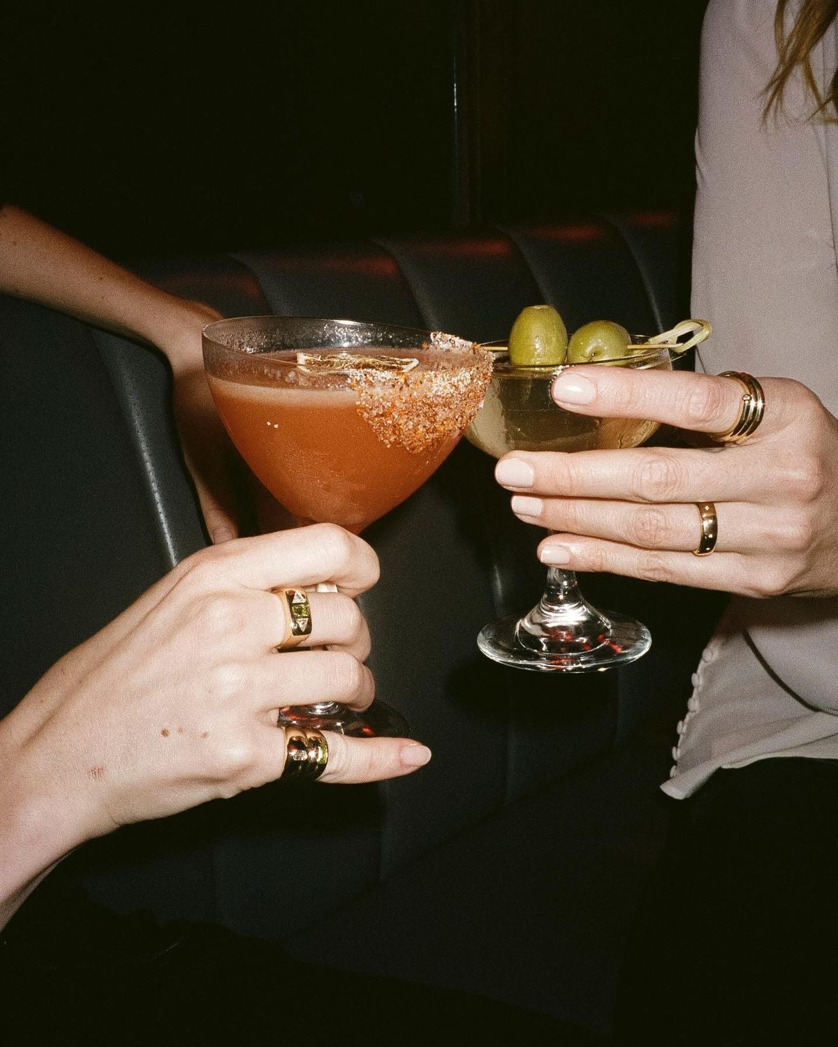 Two people holding cocktails with olives against a dark background