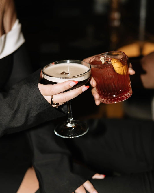 Two hands holding cocktails, one with a white foam top and the other with a red liquid and orange slice, against a blurred background.
