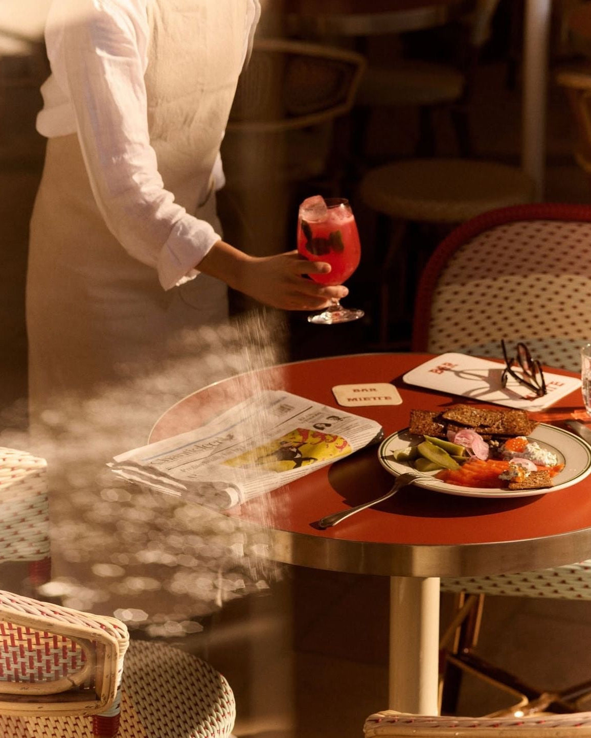 Person in a white shirt and apron holding a glass of red drink at a table with food and cutlery.